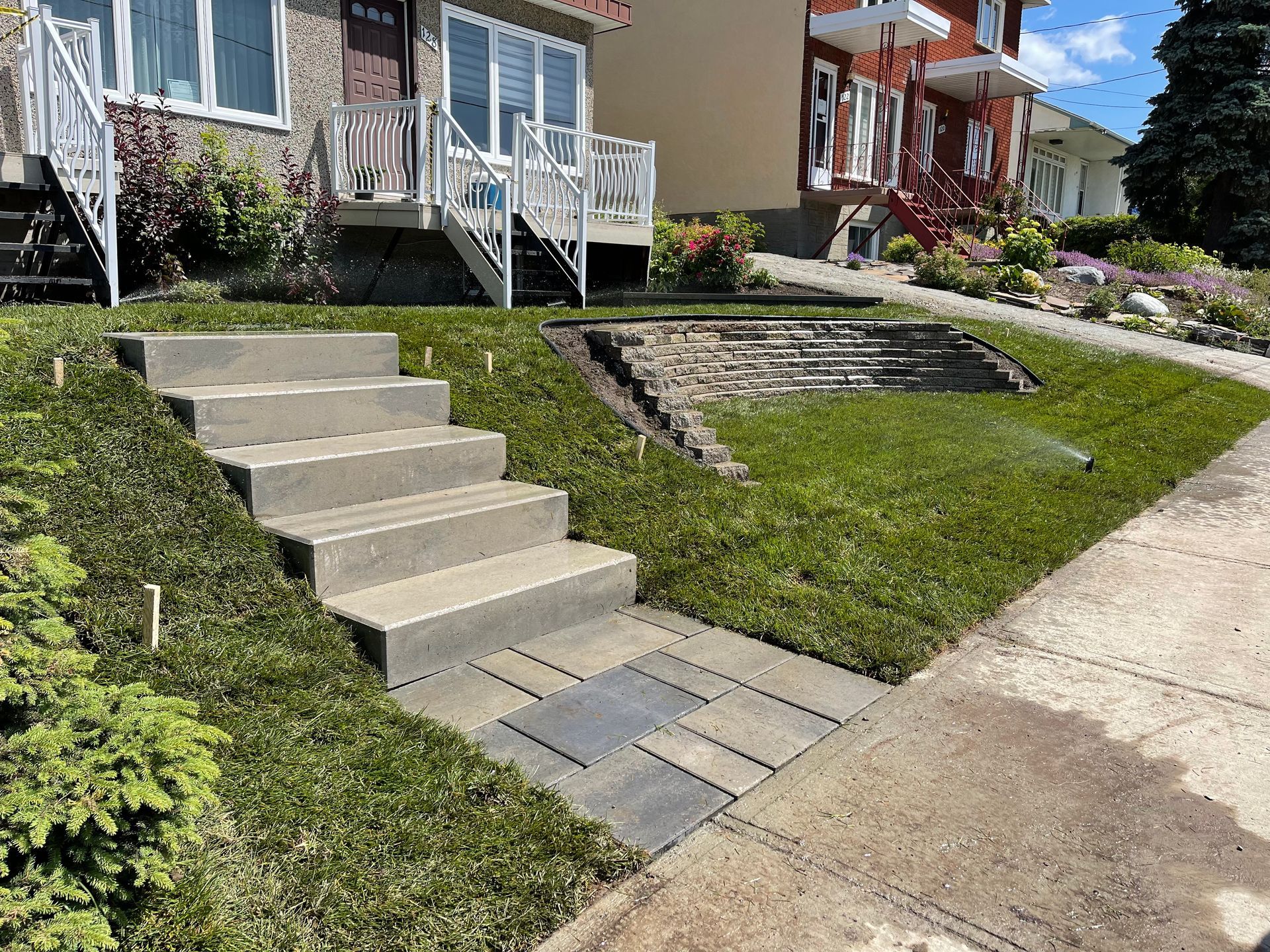 Escalier et allée en pente menant aux maisons, avec balustrades blanches et muret de soutènement engazonné.