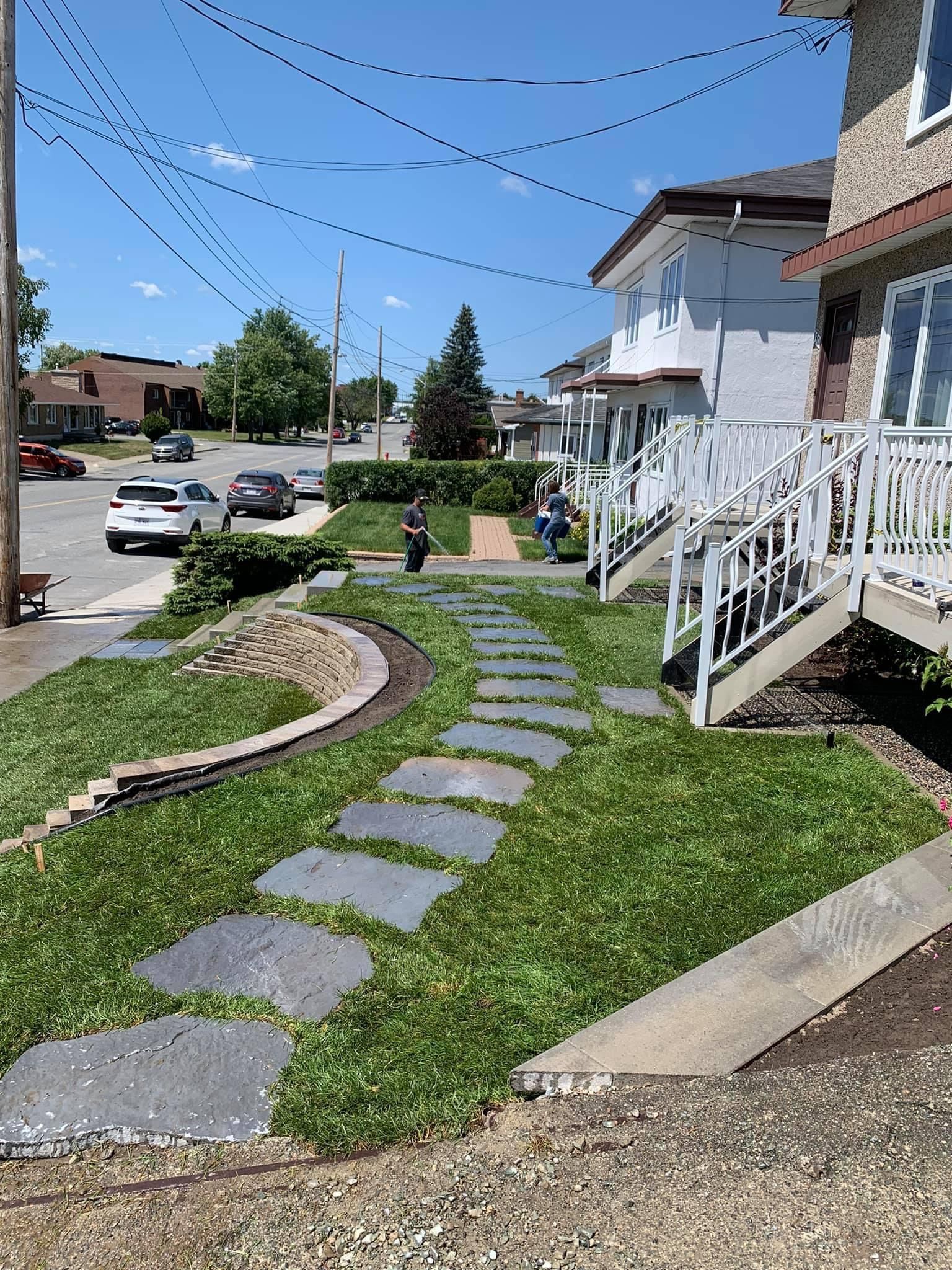 Un chemin de dalles sinueux traverse une cour avant herbeuse, à côté d'une maison blanche dans une rue ensoleillée.
