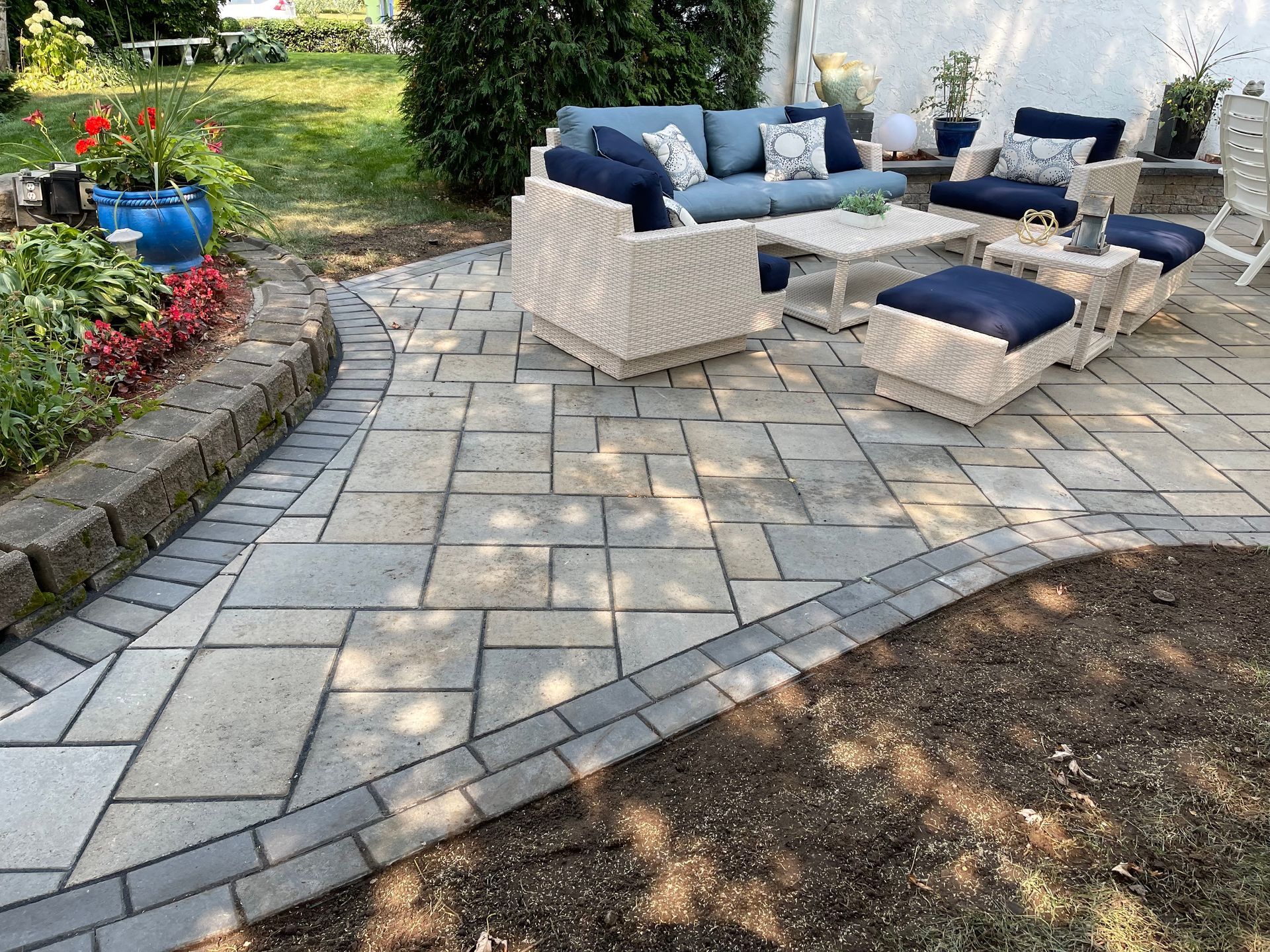 Terrasse extérieure avec dalles grises, sièges en osier, coussins bleus et petit pouf dans un cadre de jardin