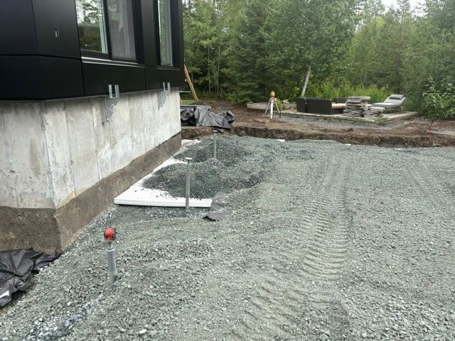 Cour recouverte de gravier à côté d'une maison en construction, avec des fondations en béton et des arbres en arrière-plan.