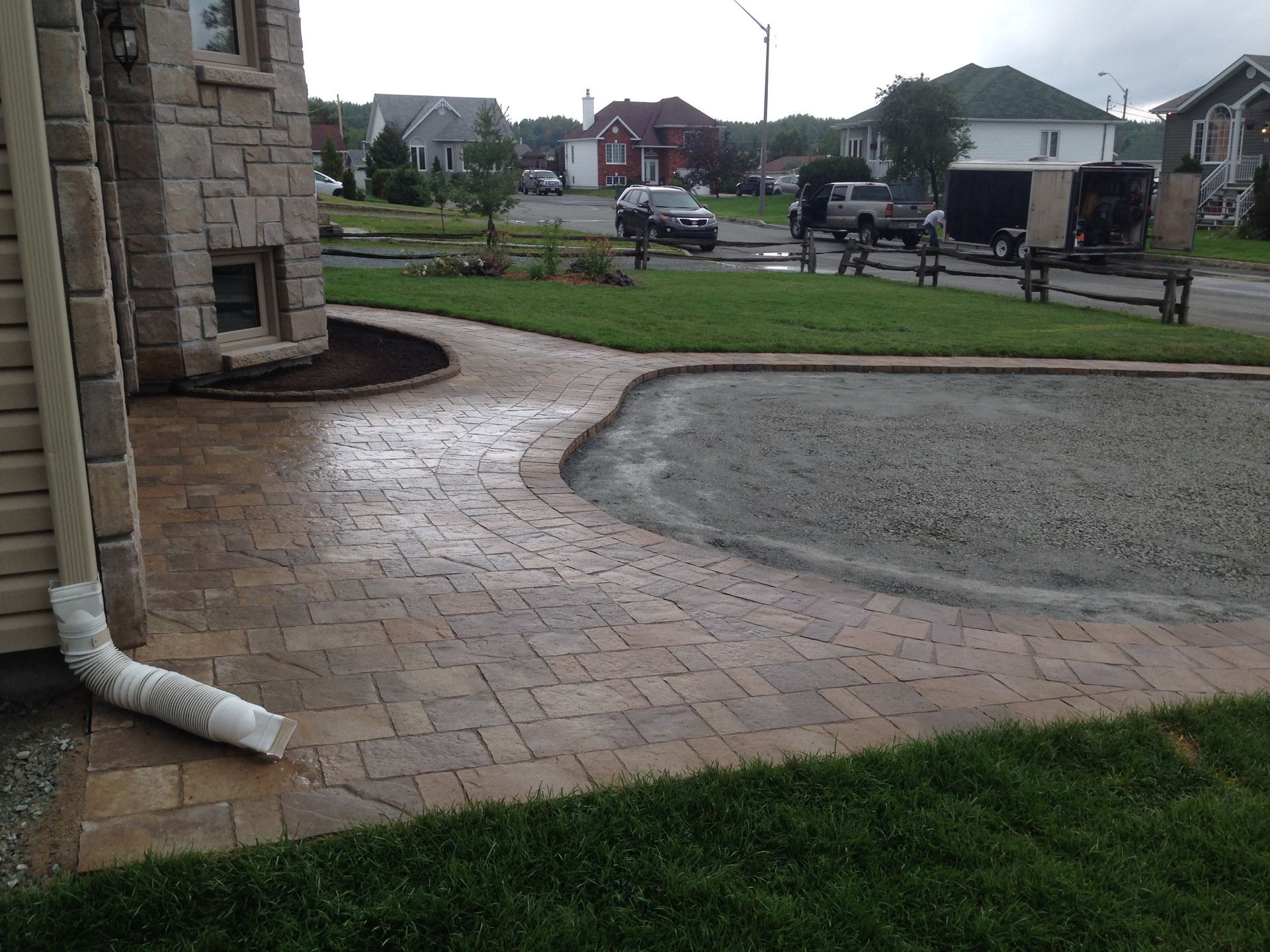 Terrasse arrière avec allée en briques, zone de gravier incurvée et un tuyau de drainage à côté d'une maison en pierre.