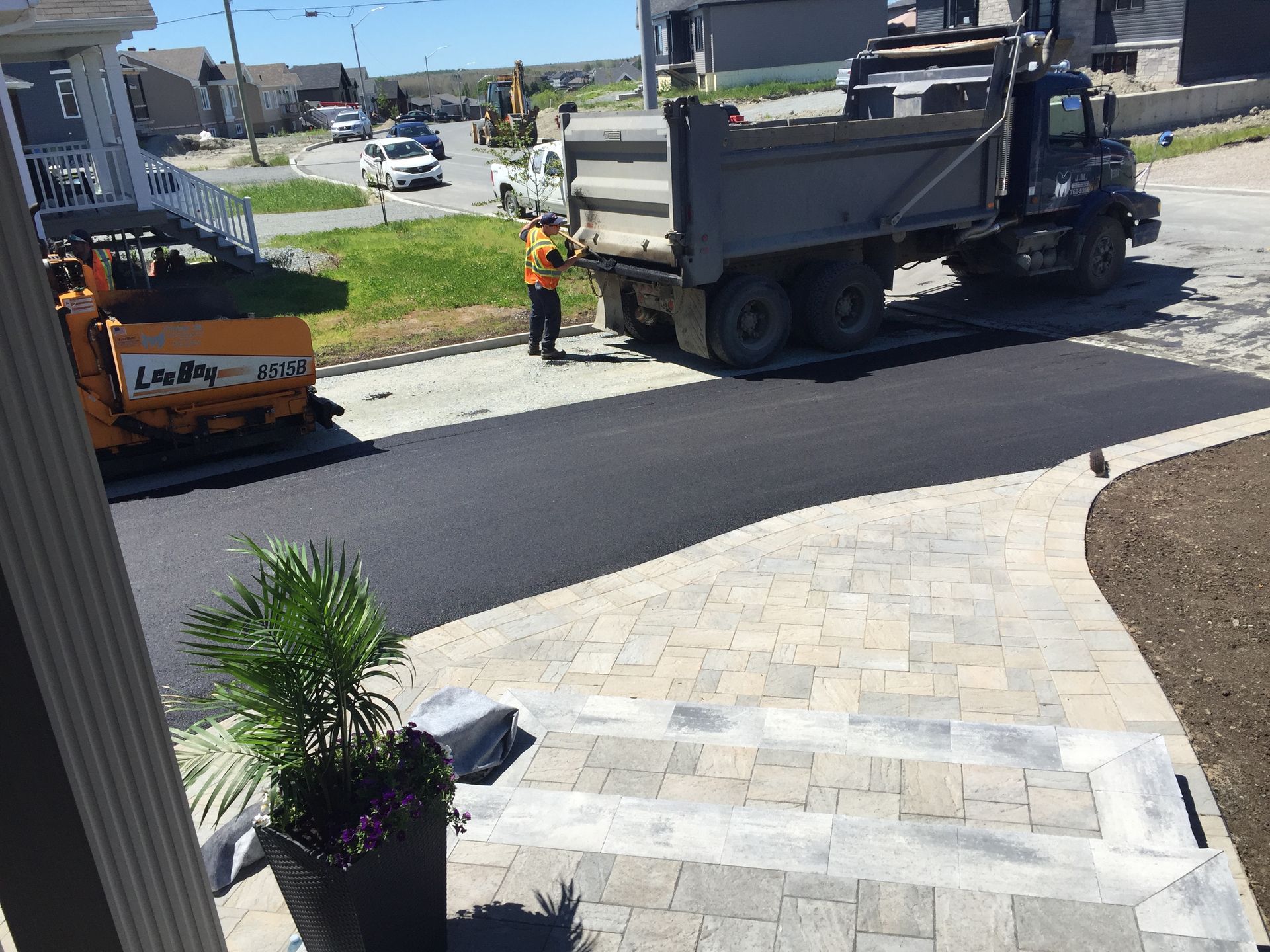 Une équipe de travaux publics paver une rue résidentielle à côté d'un camion-benne et d'une excavatrice stationnés, avec de l'asphalte noir neuf.