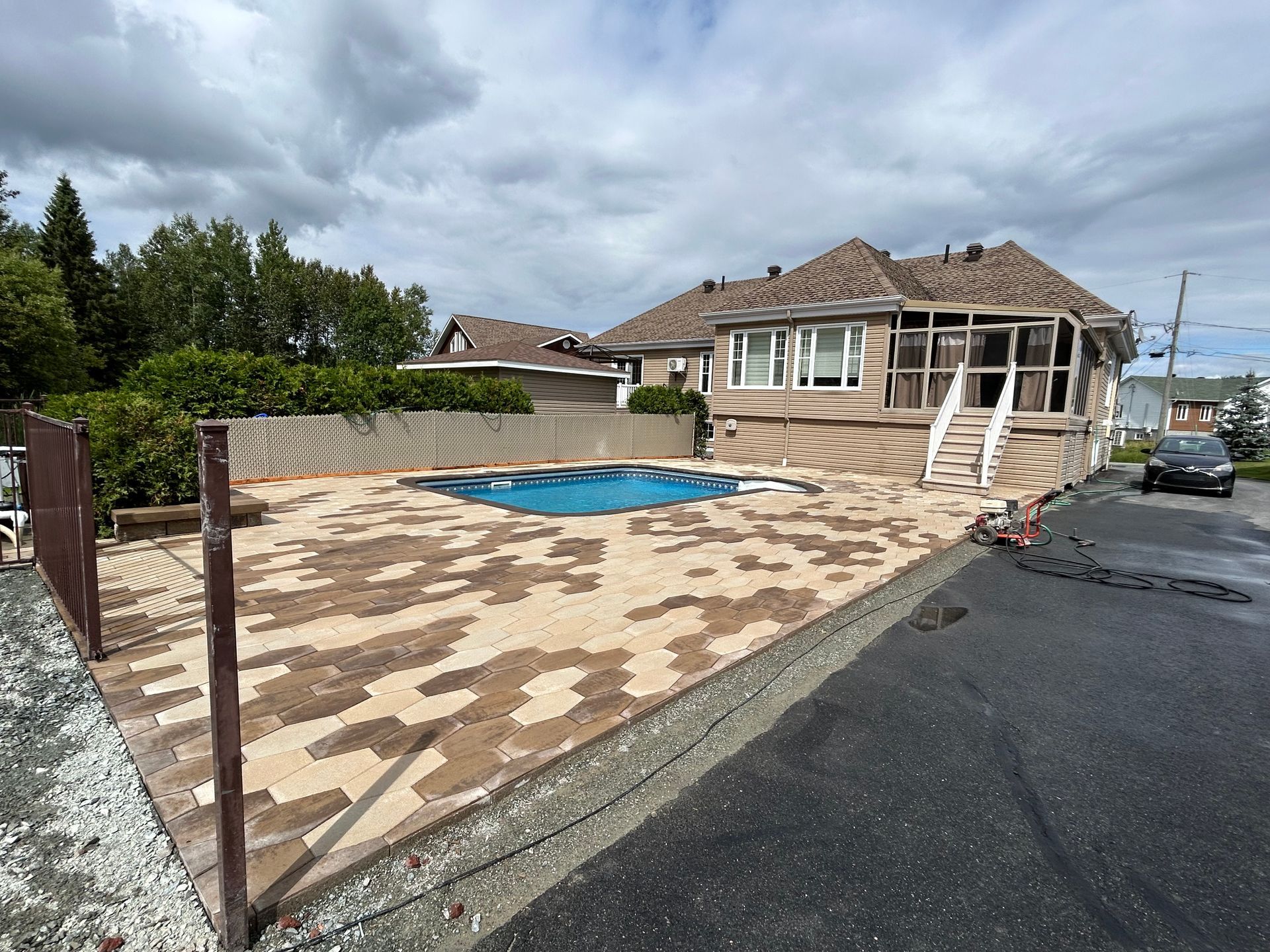 Allée résidentielle avec terrasse en pierre, petite piscine et maison beige sous un ciel nuageux.