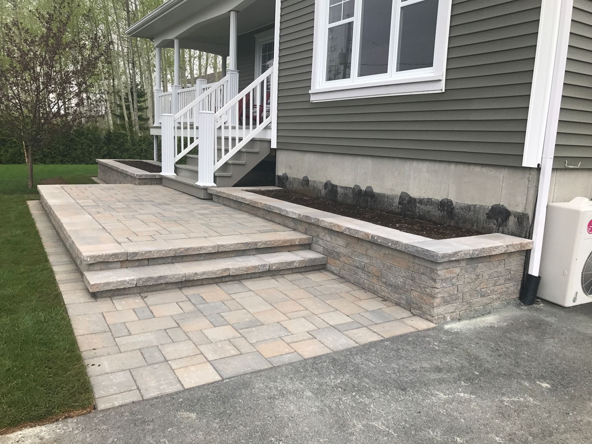 Un perron et une allée pavée mènent à une maison grise avec une balustrade blanche et une pelouse bien entretenue.