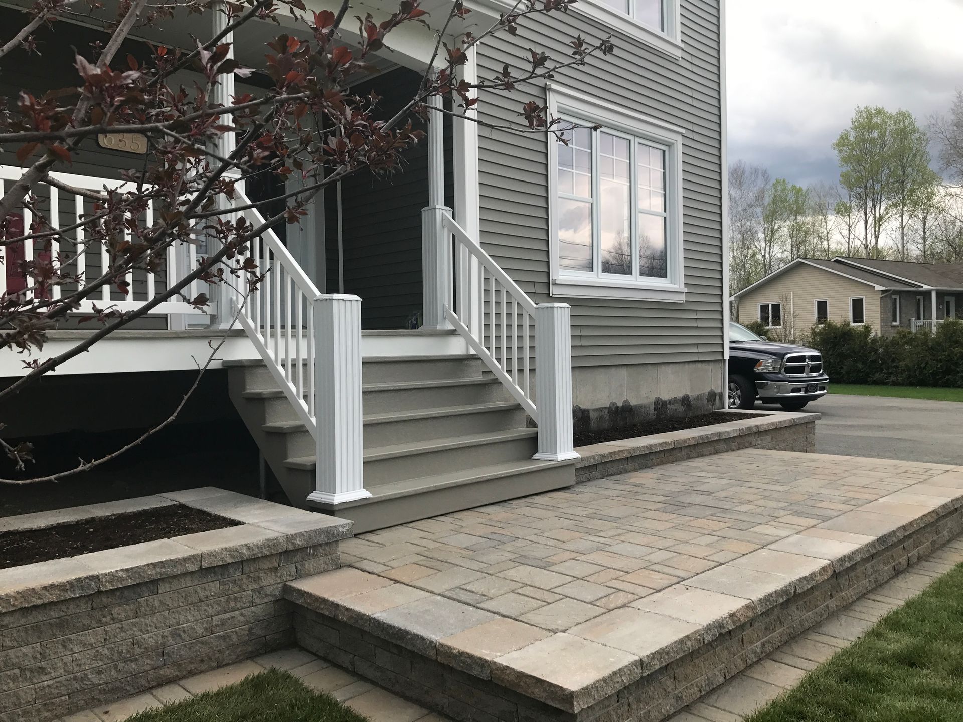 Un perron et une allée carrelée mènent à l'entrée d'une maison avec une balustrade blanche et une voiture garée à proximité.