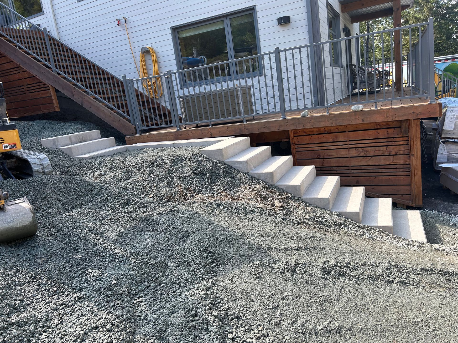 Cour arrière avec pente de gravier, mur de soutènement en bois, escalier et terrasse avec rambarde.