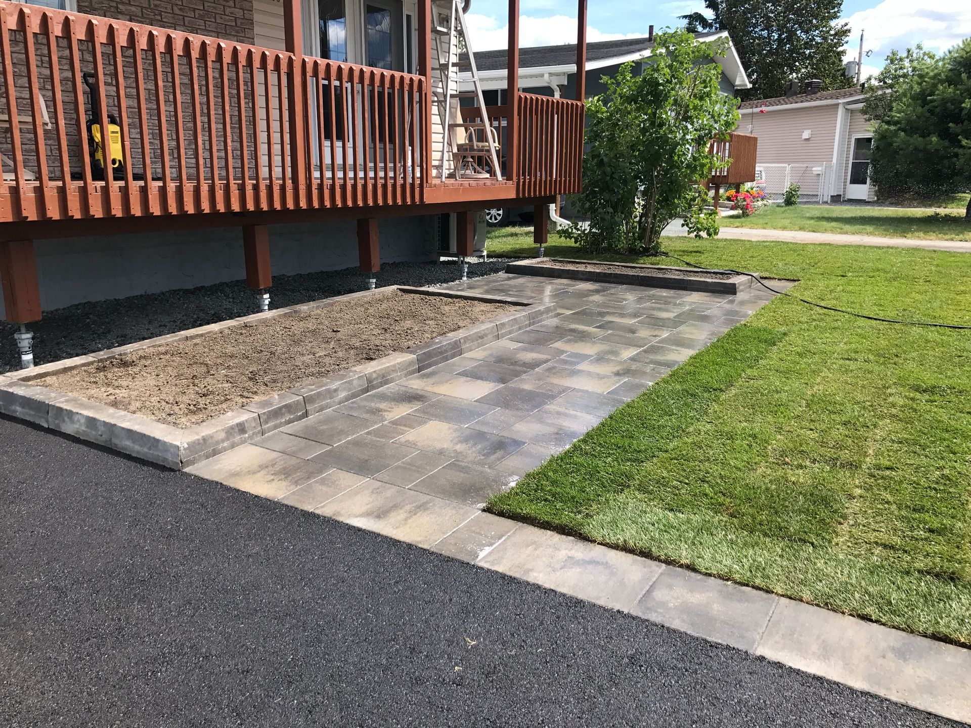 Terrasse d'appartement avec plancher en bois, pavés de pierre et pelouse adjacente à une allée asphaltée.