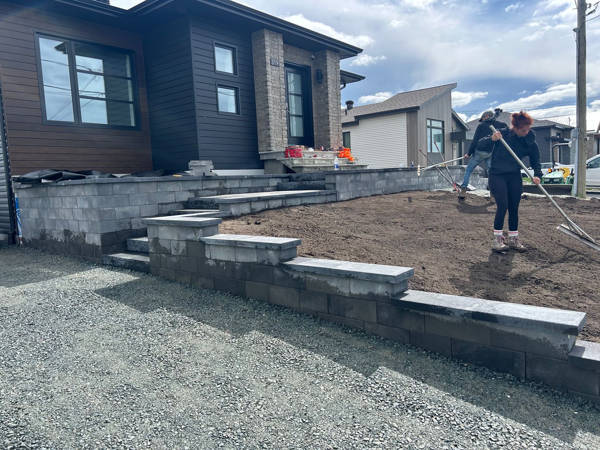 Cour résidentielle en construction avec des marches en pierre, du gravier et un ouvrier utilisant une pelle près d'une maison