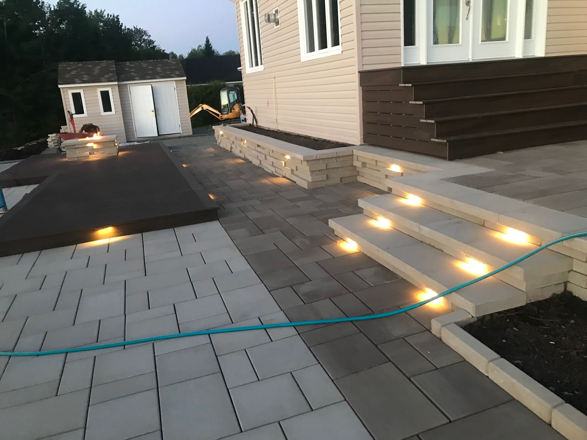 Terrasse extérieure avec marches éclairées, dalles de pierre et tuyau d'arrosage turquoise près d'une maison au crépuscule