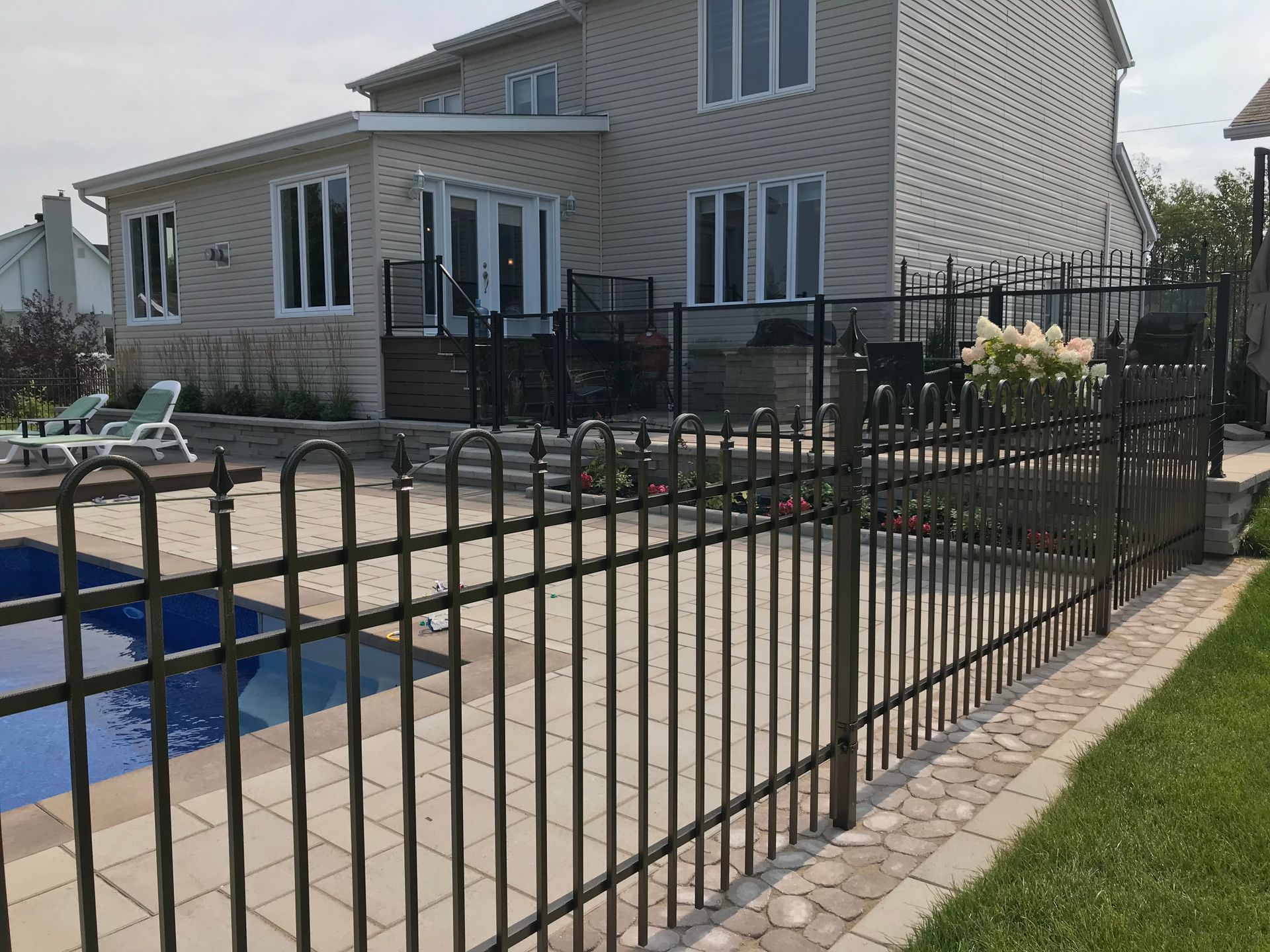 Terrasse arrière avec clôture en métal noir, piscine, chaises longues et maison beige.