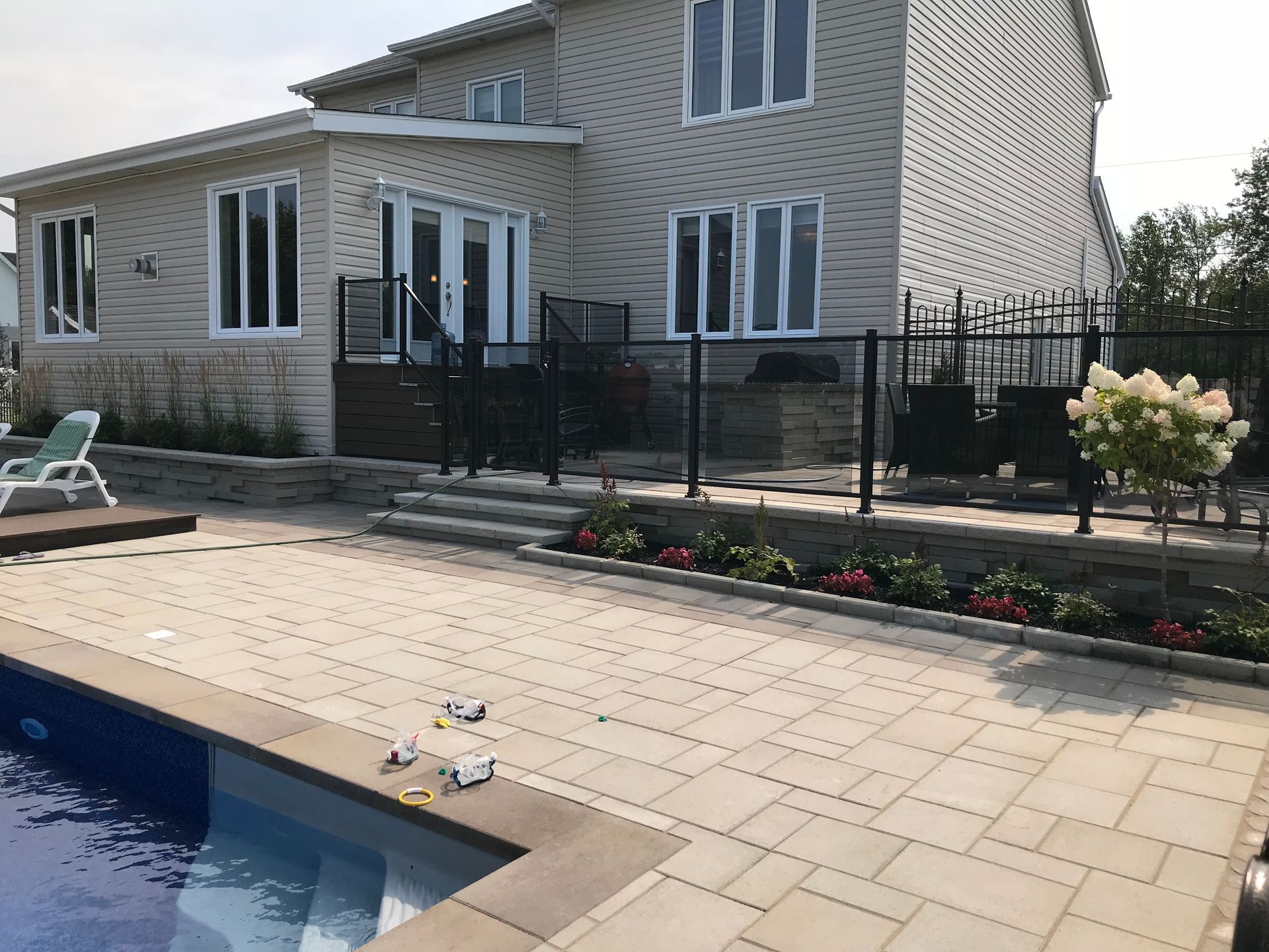 Terrasse arrière donnant sur une piscine et une maison beige avec une balustrade noire et des parterres de fleurs