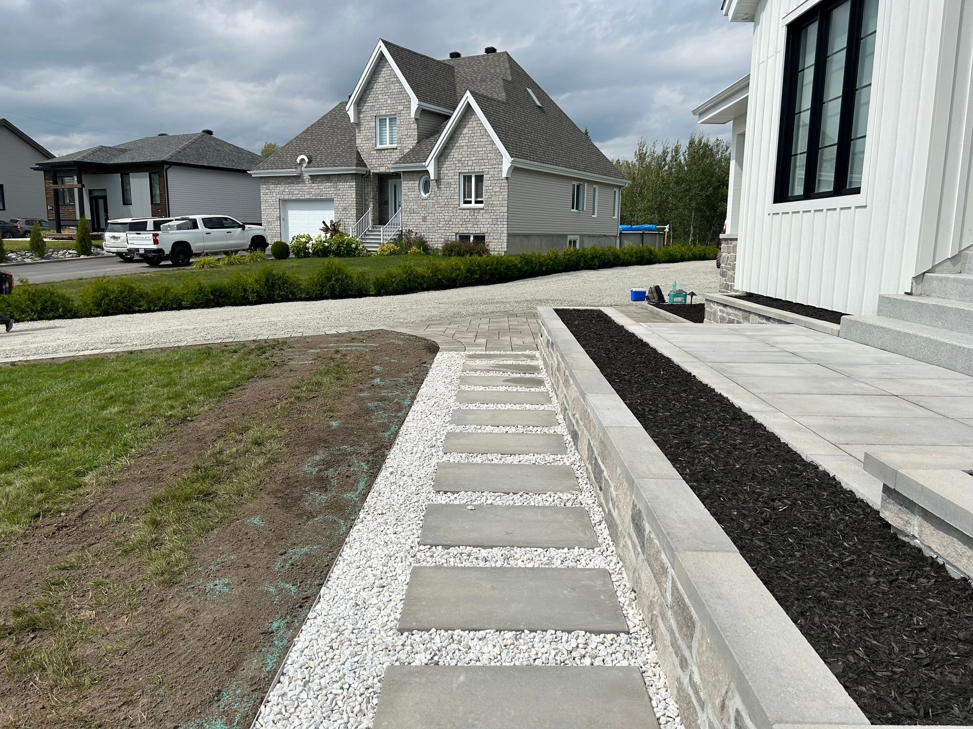 Allée pavée de dalles grises dans la cour avant d'une maison blanche, menant à une rue résidentielle bordée de maisons et de pelouses.
