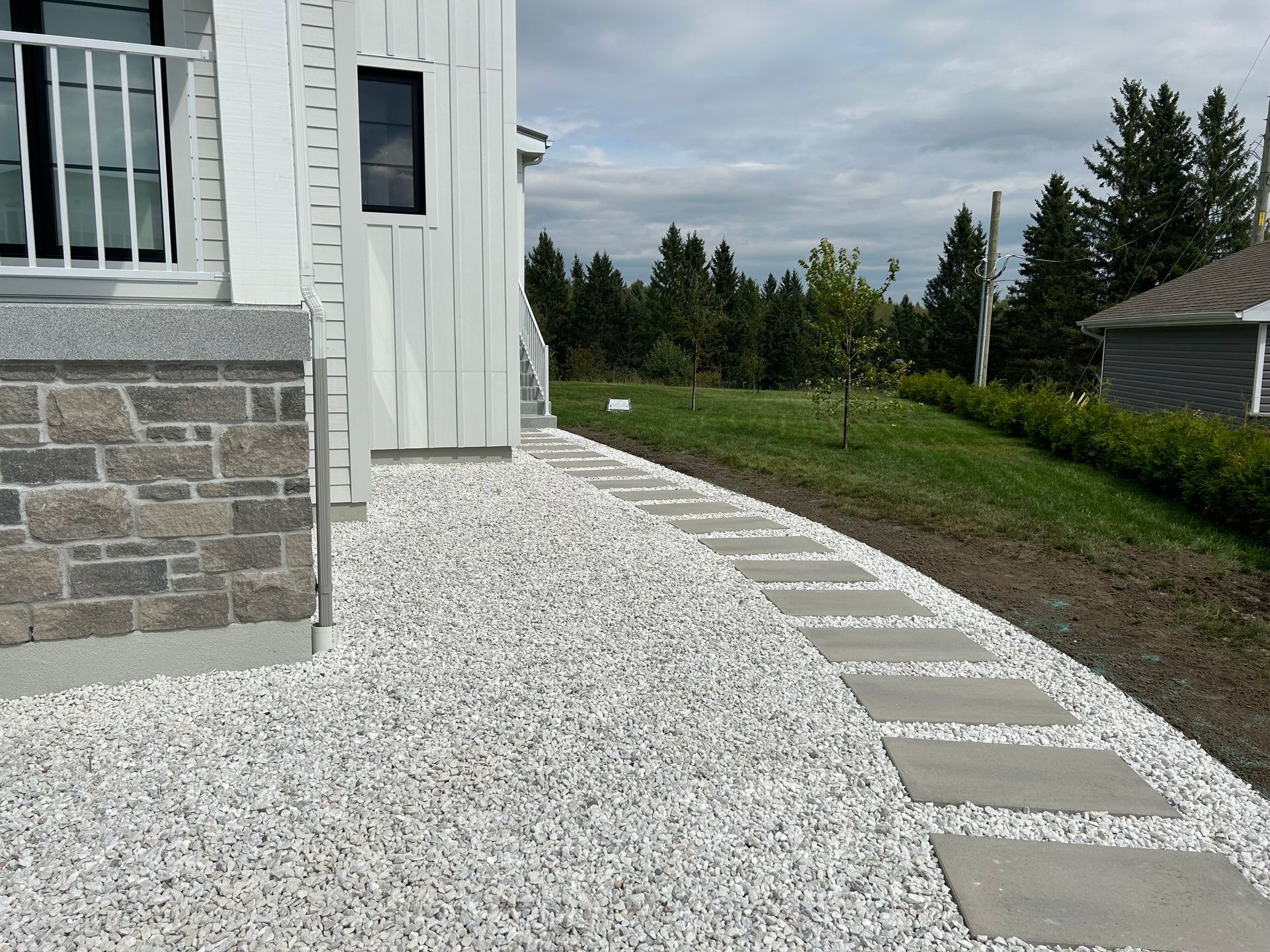 Allée de gravier blanc longeant une maison, avec des pas japonais menant à une cour verdoyante bordée d'arbres.