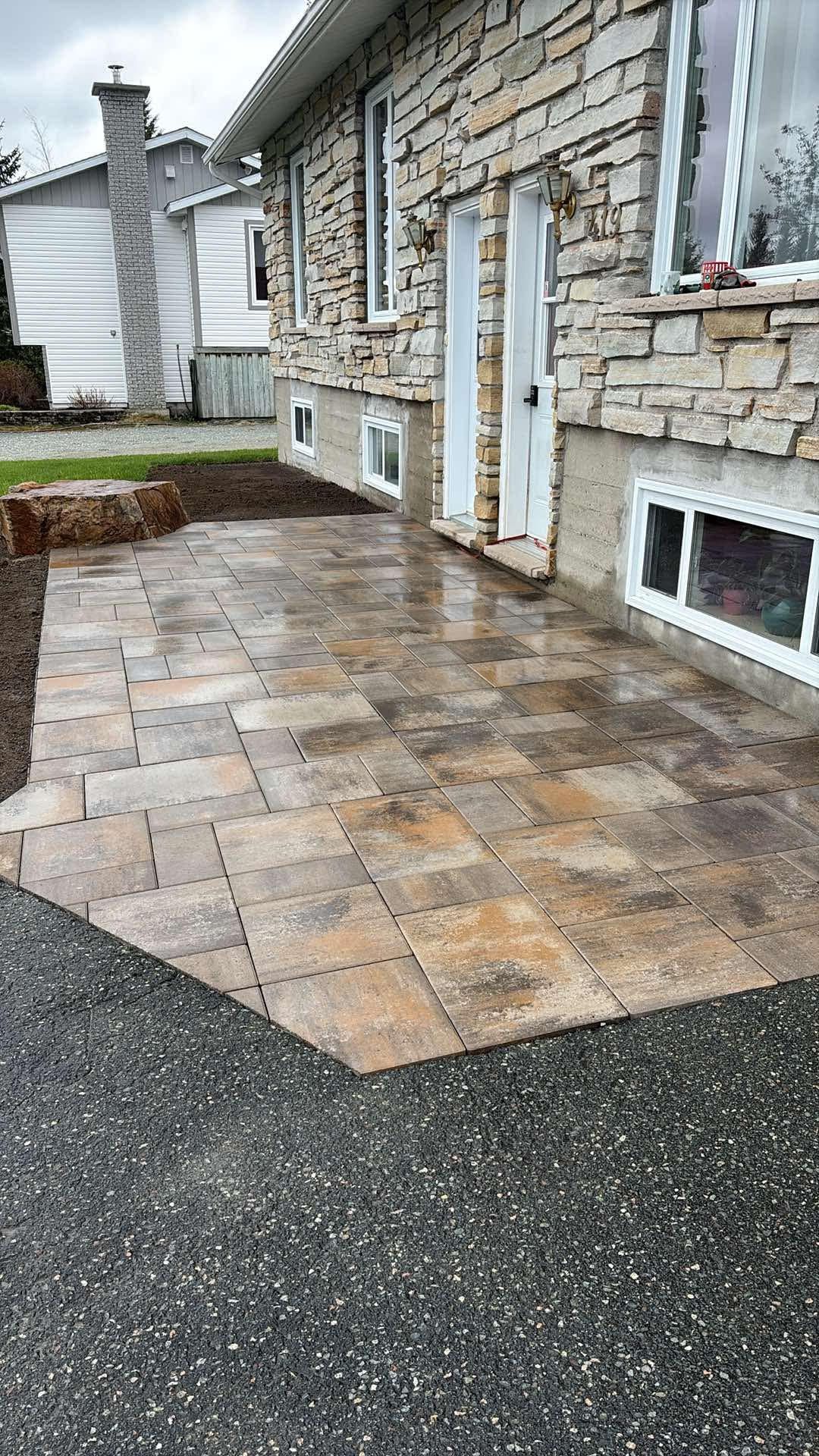 Terrasse en pierre jouxtant une maison en briques, bordée par une allée en asphalte foncé.