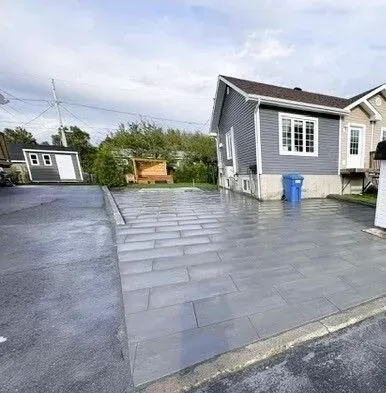 Allée pavée grise à côté d'une petite maison, avec une poubelle bleue et un banc en bois dans la cour