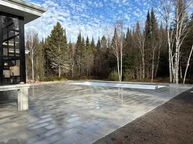 Terrasse extérieure en pierre jouxtant un bâtiment moderne, bordée d'arbres sous un ciel partiellement nuageux.