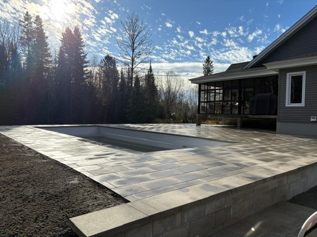 Terrasse arrière partiellement pavée à côté d'une maison, avec un mur de soutènement en pierre ensoleillé et des arbres en arrière-plan