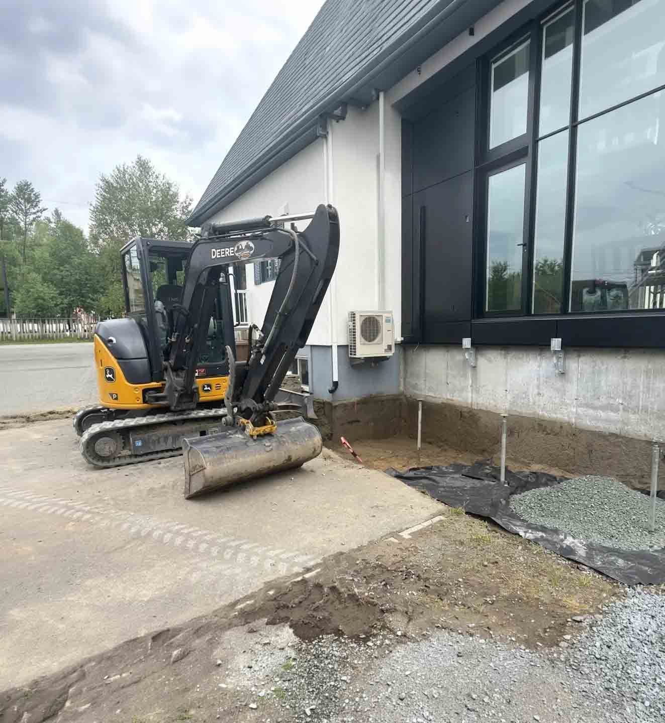 Une pelleteuse jaune se trouve à côté d'un bâtiment, dégageant la terre et le gravier près d'un mur et d'une fenêtre.