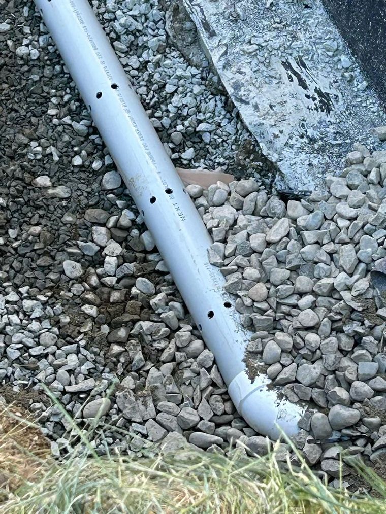 Un tuyau de drainage blanc perforé repose sur du gravier, à côté d'un trottoir en béton et de gazon.