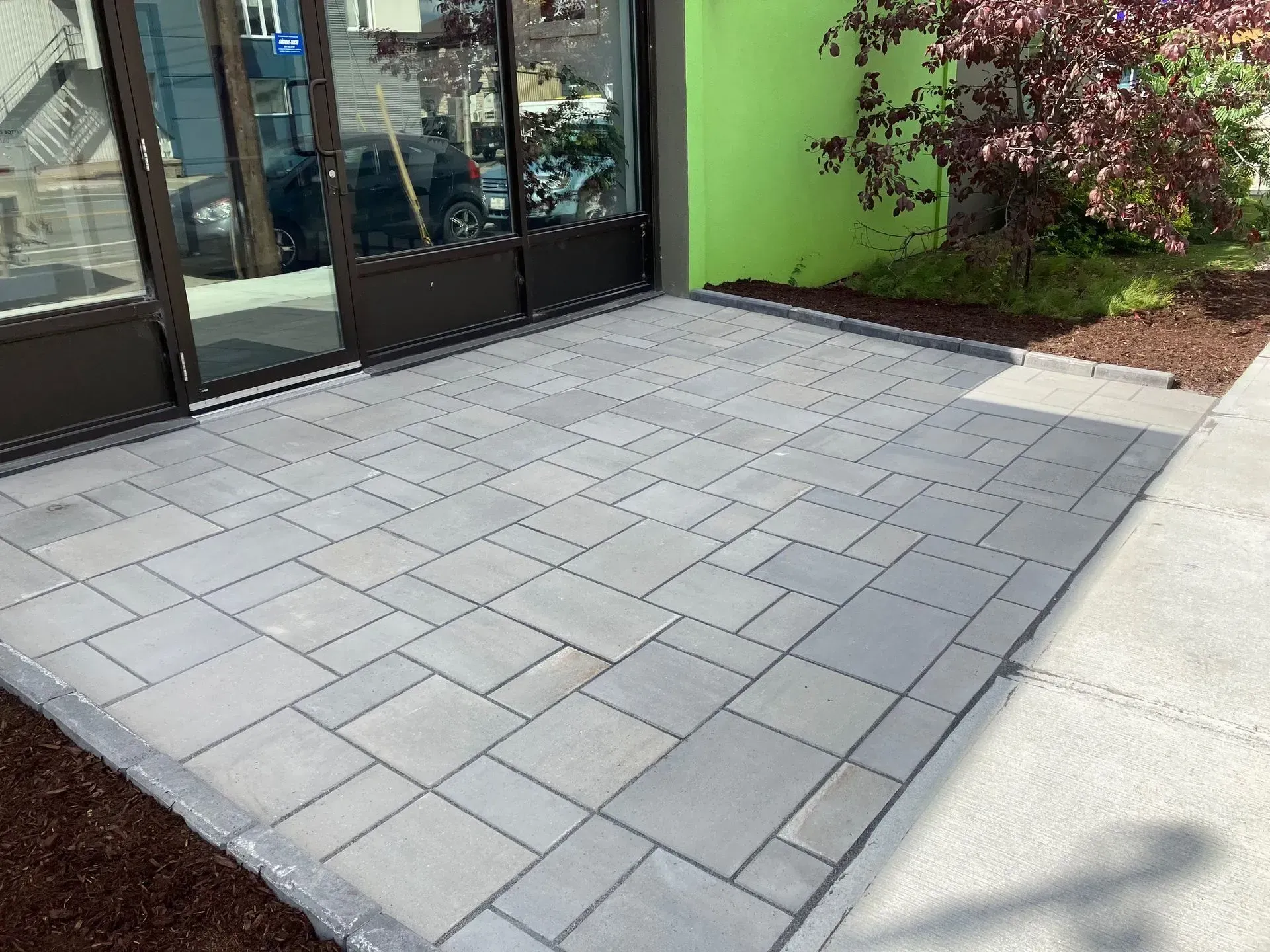 Terrasse pavée de dalles gris clair jouxtant une vitrine et un mur végétal, avec un arbuste dans un coin.