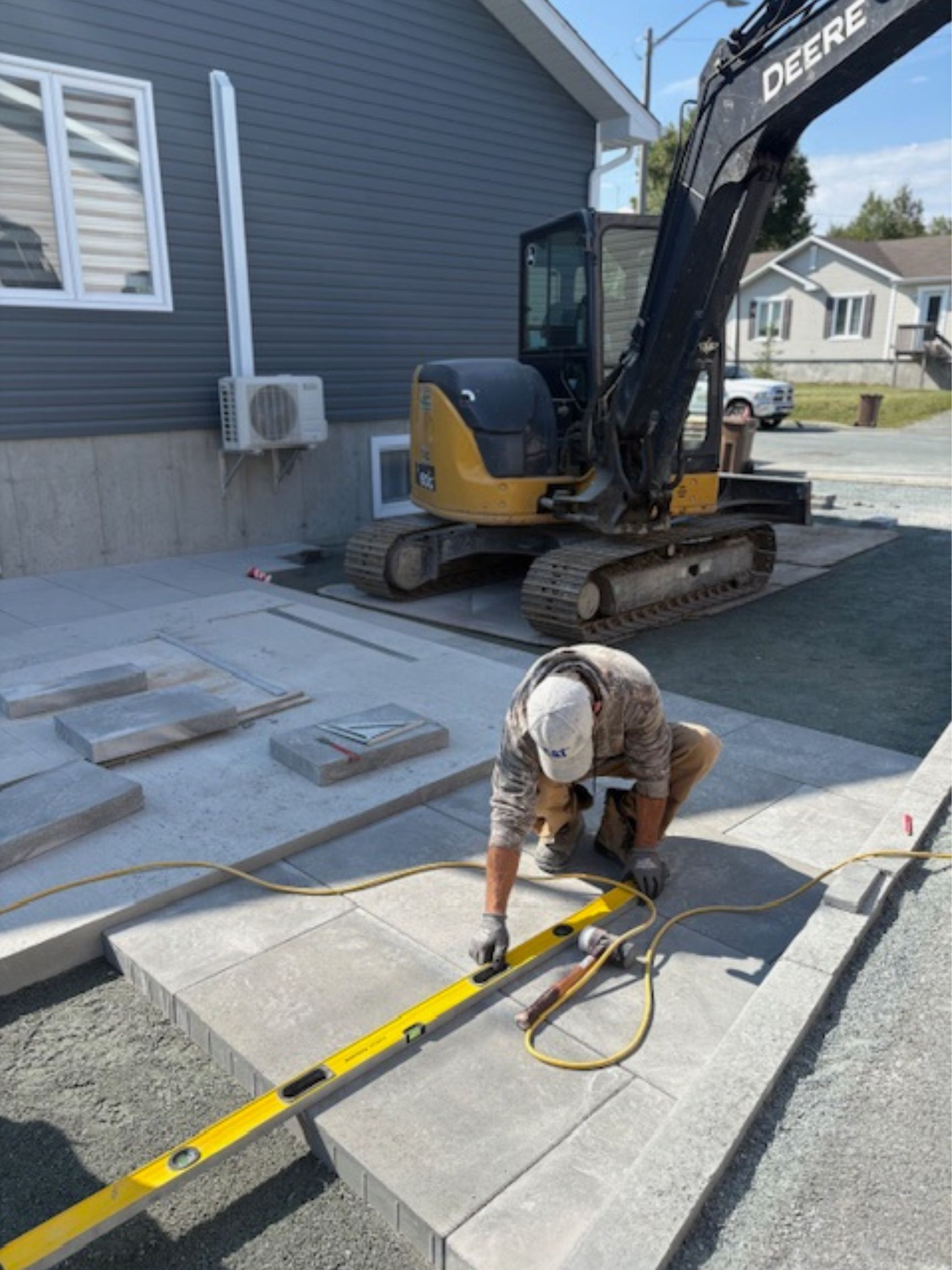 Un ouvrier mesure des dalles de béton à côté d'une mini-pelle sur une allée.