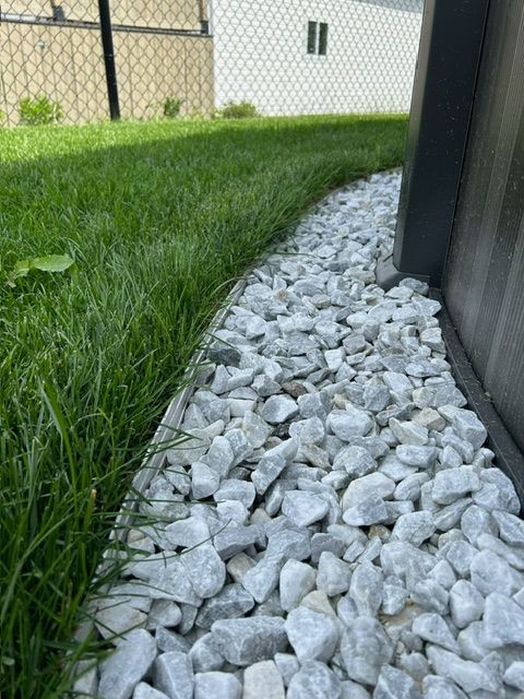 De l'herbe borde une bande de drainage en pierre blanche le long d'un mur d'immeuble, avec une clôture en grillage en arrière-plan.