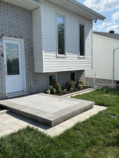 Extérieur de la maison avec une terrasse avant surélevée en béton, un revêtement blanc et de petits parterres de jardin.