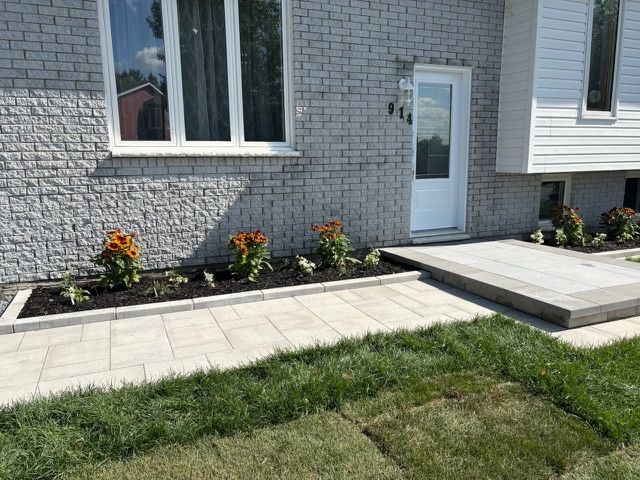 Façade d'une maison en briques grises avec une porte blanche, des fenêtres, un petit parterre de fleurs et une allée pavée.