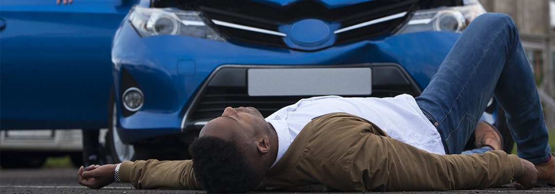A man is laying on the ground in front of a blue car. A man is laying on the ground in front of a blue car.