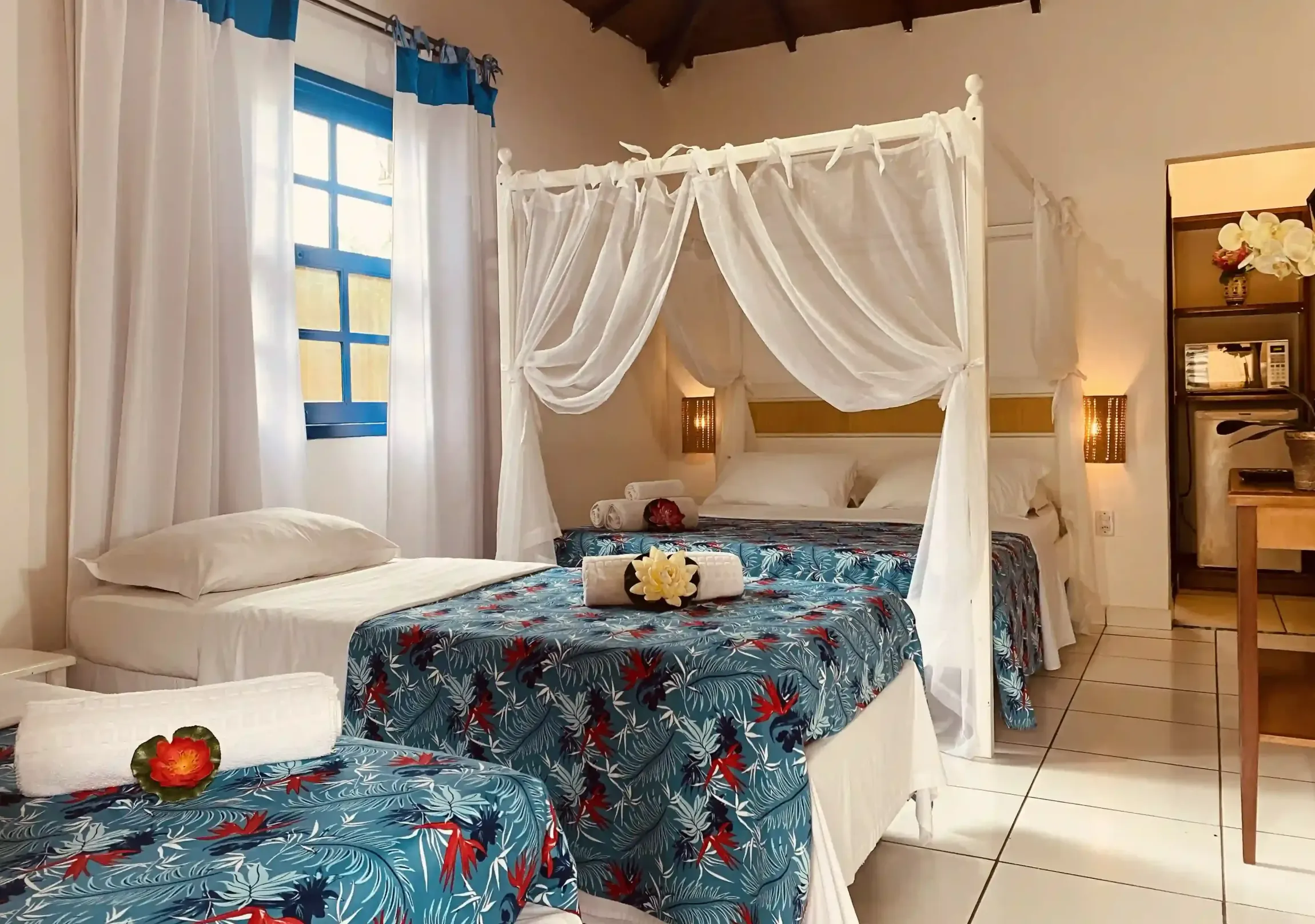 Quarto luminoso com duas camas, cobertas com estampa floral azul e um dossel branco sobre a cama maior.