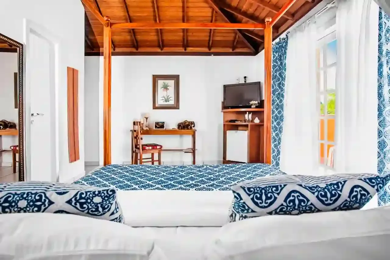 Sala de estar iluminada com sofá branco, almofadas estampadas em azul, teto de madeira, escrivaninha, TV e cortinas abertas.