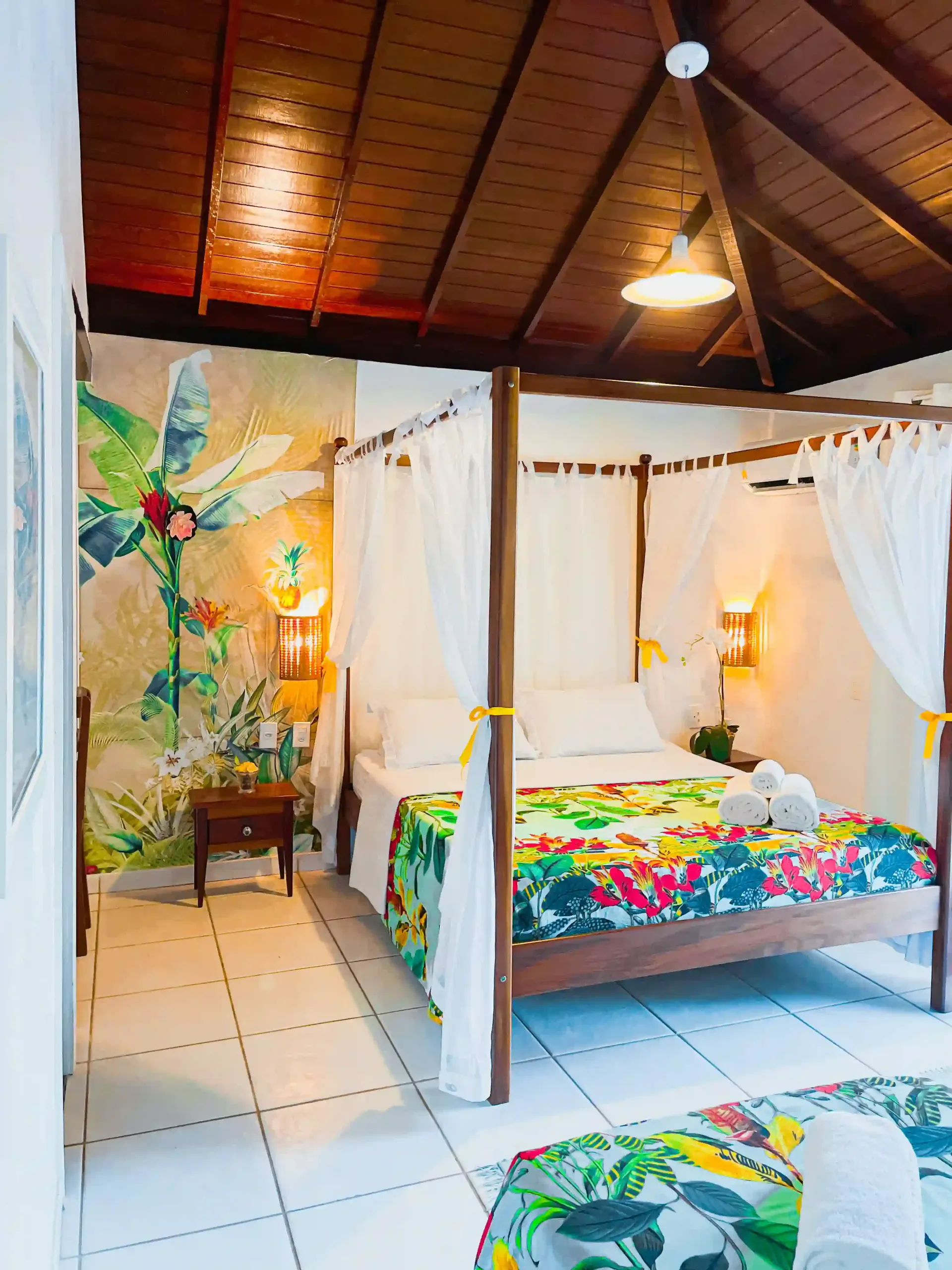 Quarto tropical aconchegante com cama de dossel branca, colcha colorida, piso de azulejo e teto abobadado de madeira.