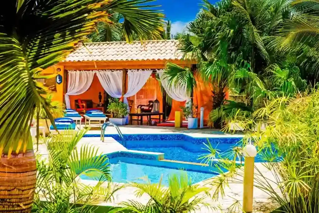 Piscina de resort tropical com água azul, espreguiçadeiras e um pátio sombreado rodeado por palmeiras.