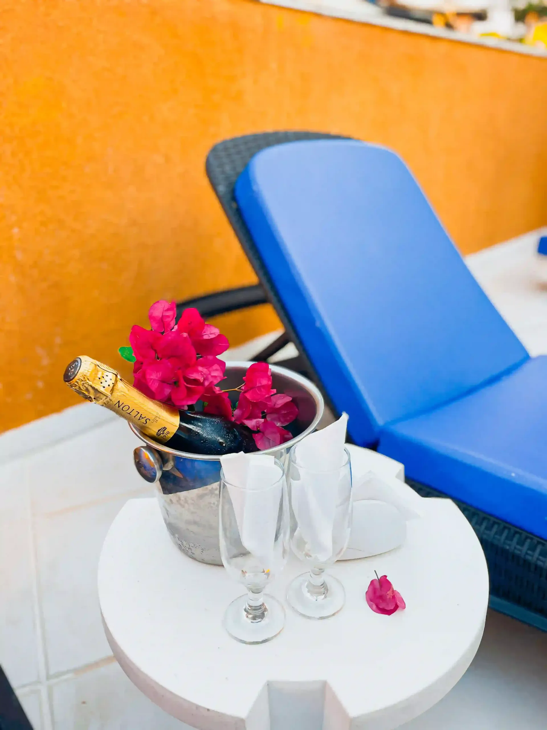 Poltrona azul ao lado de uma mesa branca com champanhe, taças e flores cor-de-rosa contra uma parede laranja.