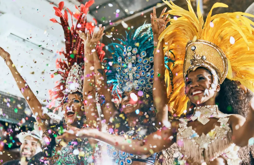 Dançarinos de carnaval com cocares de penas coloridos lançando confetes durante um desfile.