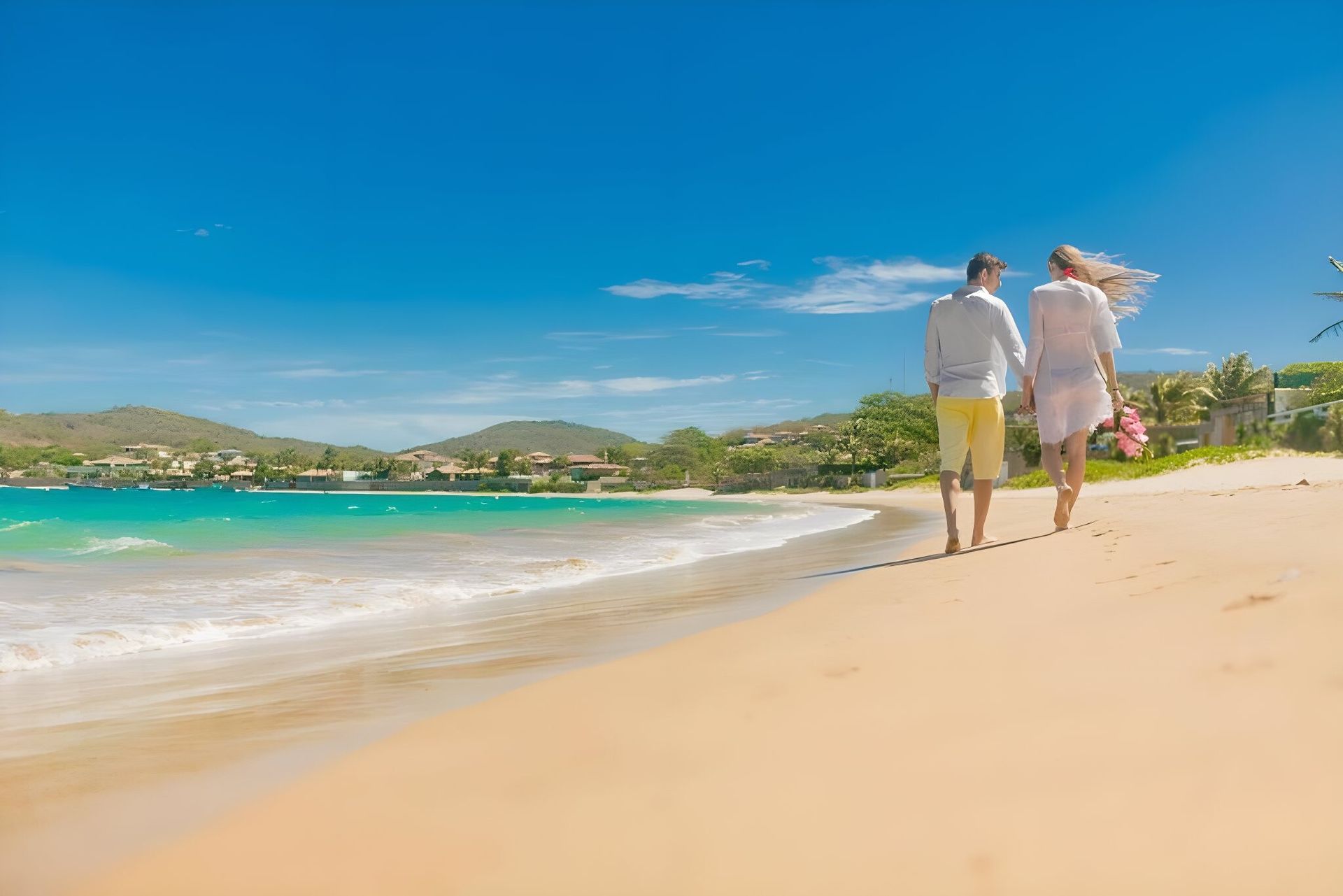 Casal caminhando por uma praia tropical ensolarada com água turquesa e colinas ao longe.