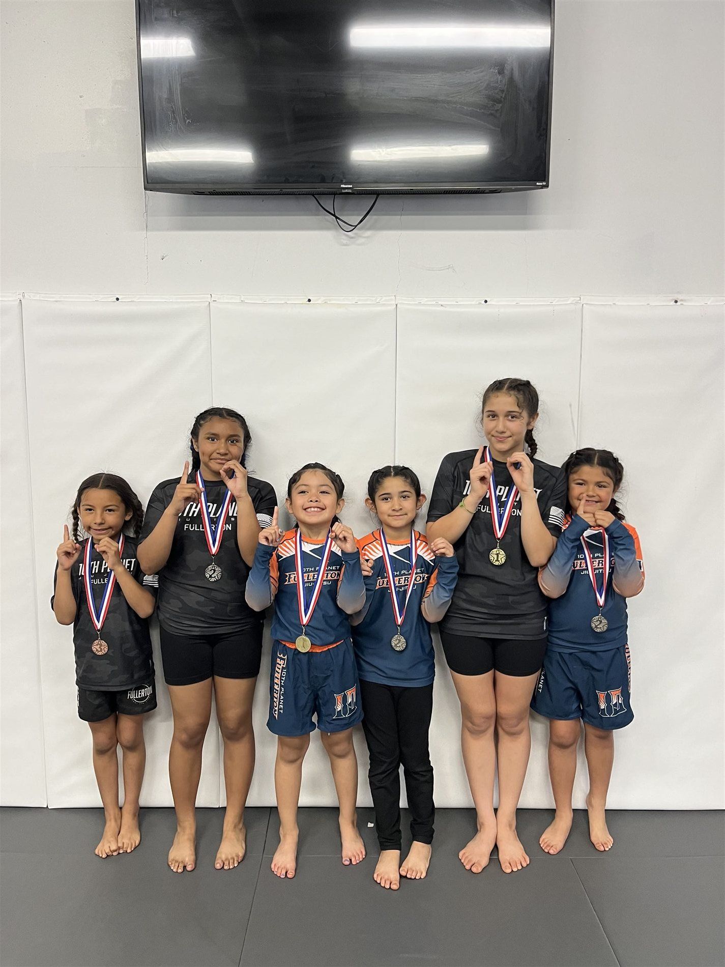 Six young athletes wearing medals pose indoors beneath a TV after a competition.