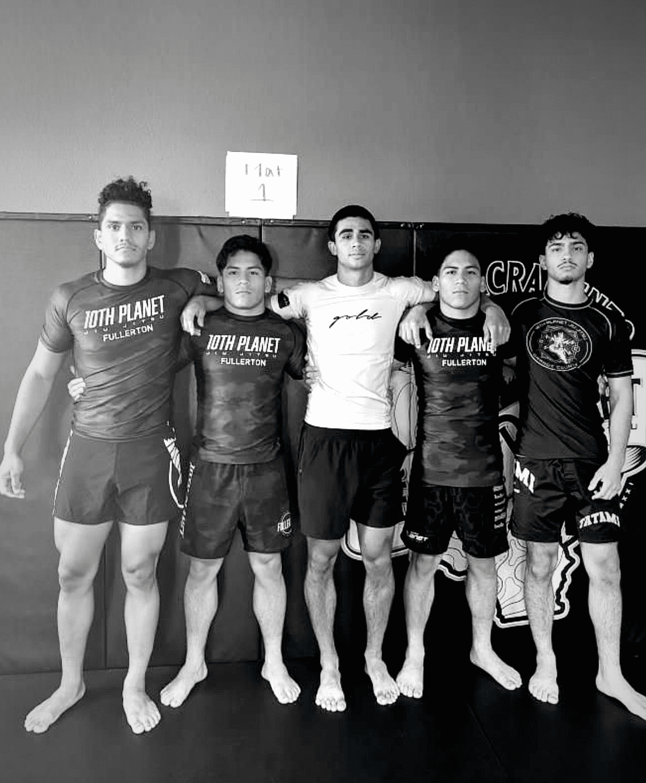 Five barefoot athletes pose in a row indoors, standing against a wall in training gear.