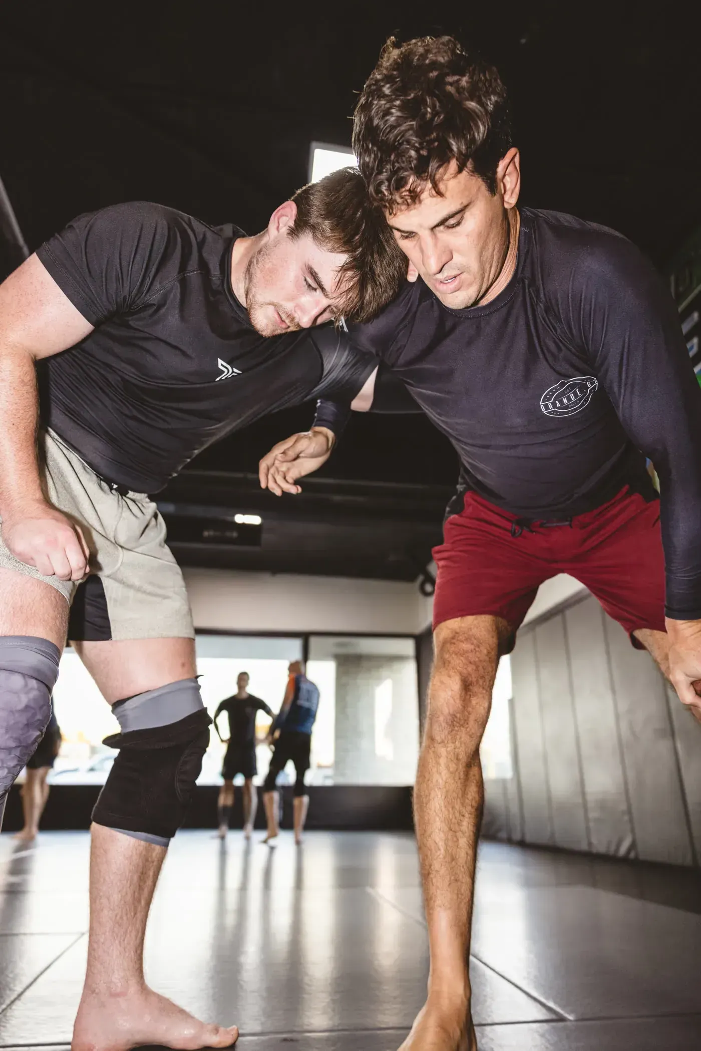 Two wrestlers in a gym, bent forward in a grapple stance, wearing training clothes.