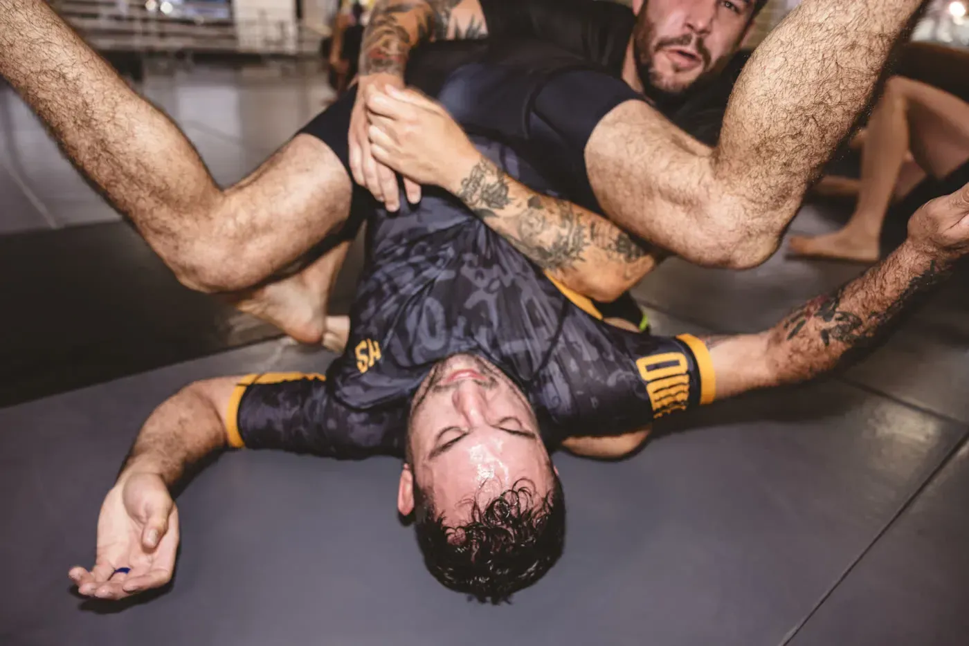 Two wrestlers grappling on a gym mat, one pinned on his back with legs around the other.