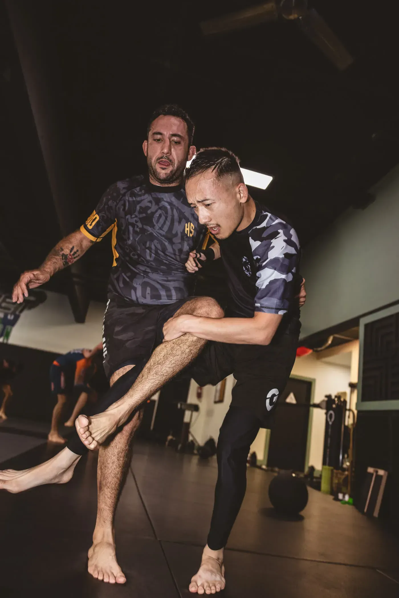 Two athletes practicing a balance drill in a gym, one supporting the other’s extended leg