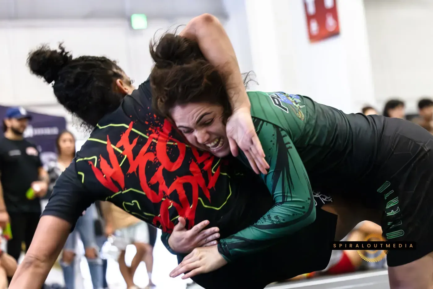 Two wrestlers grappling on a mat, one in black and red, the other in green, in a crowded gymnasium.