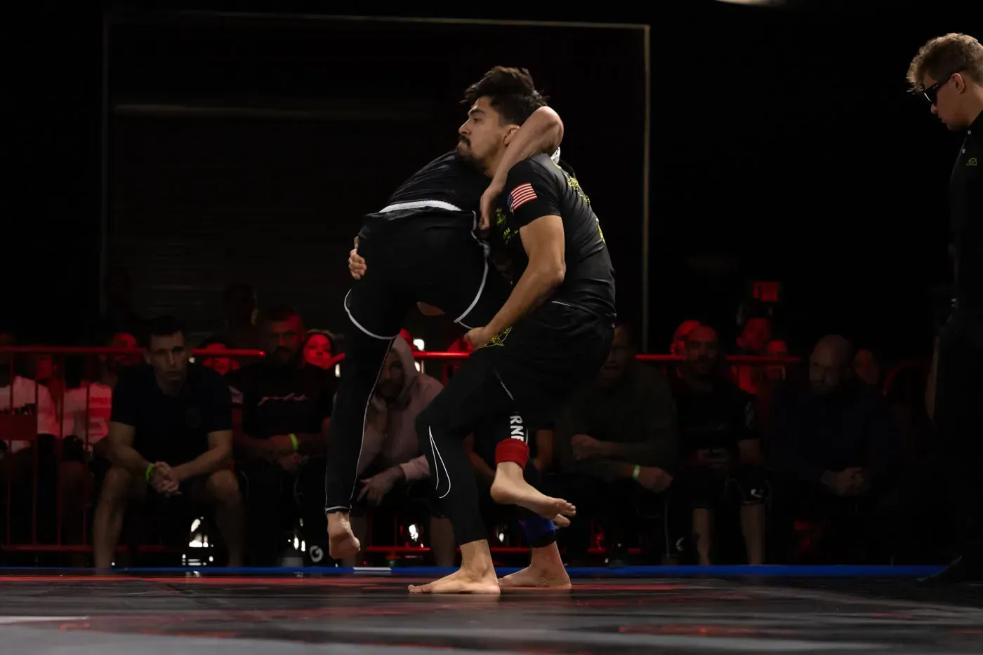 Two martial artists grapple on a lit stage, one lifting the other, with spectators in the background.