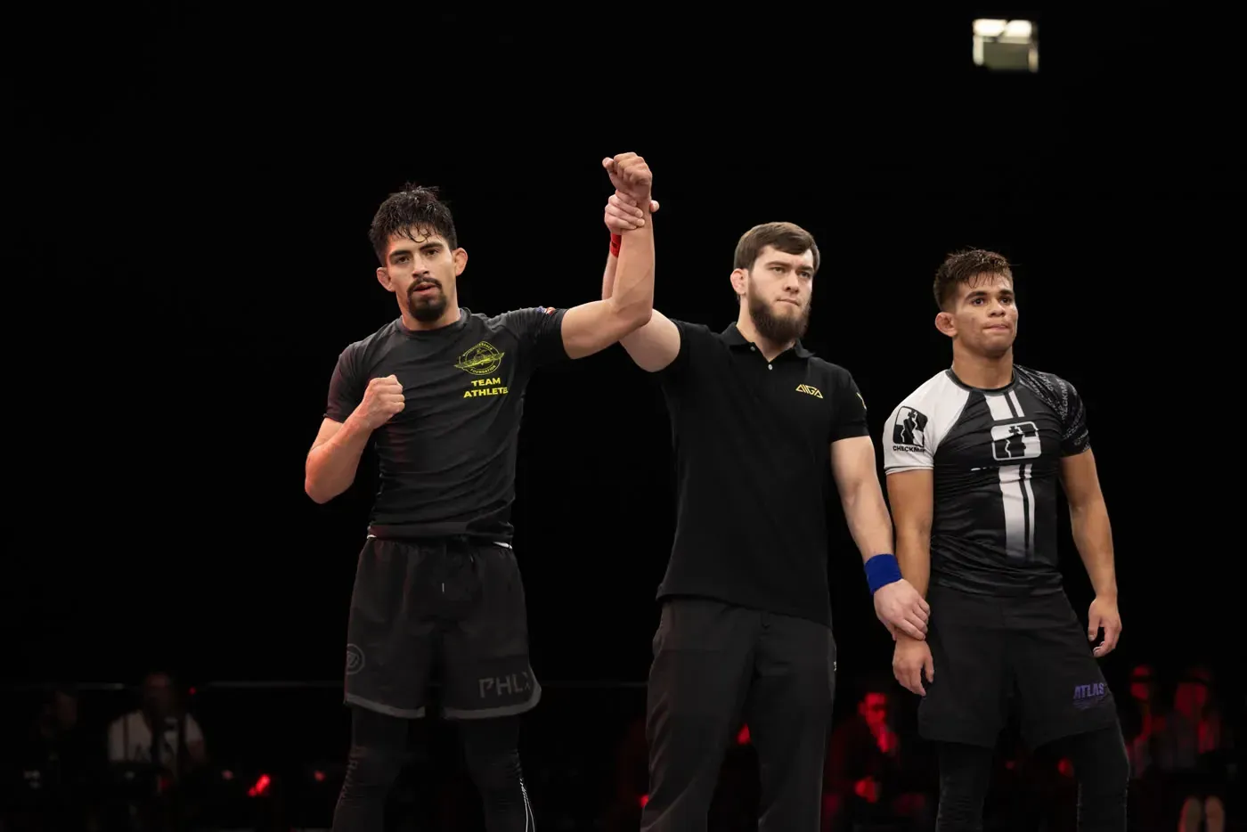 Three MMA fighters stand in the ring as the referee raises the winner’s arm.