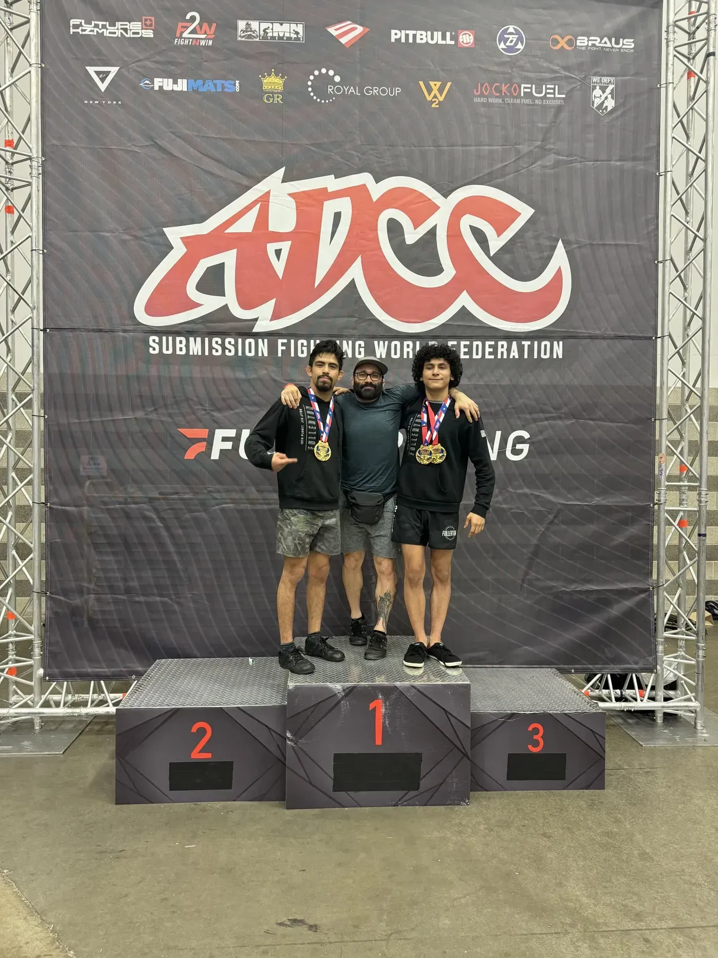 Three athletes on a podium with medals in front of an ADCC championship backdrop