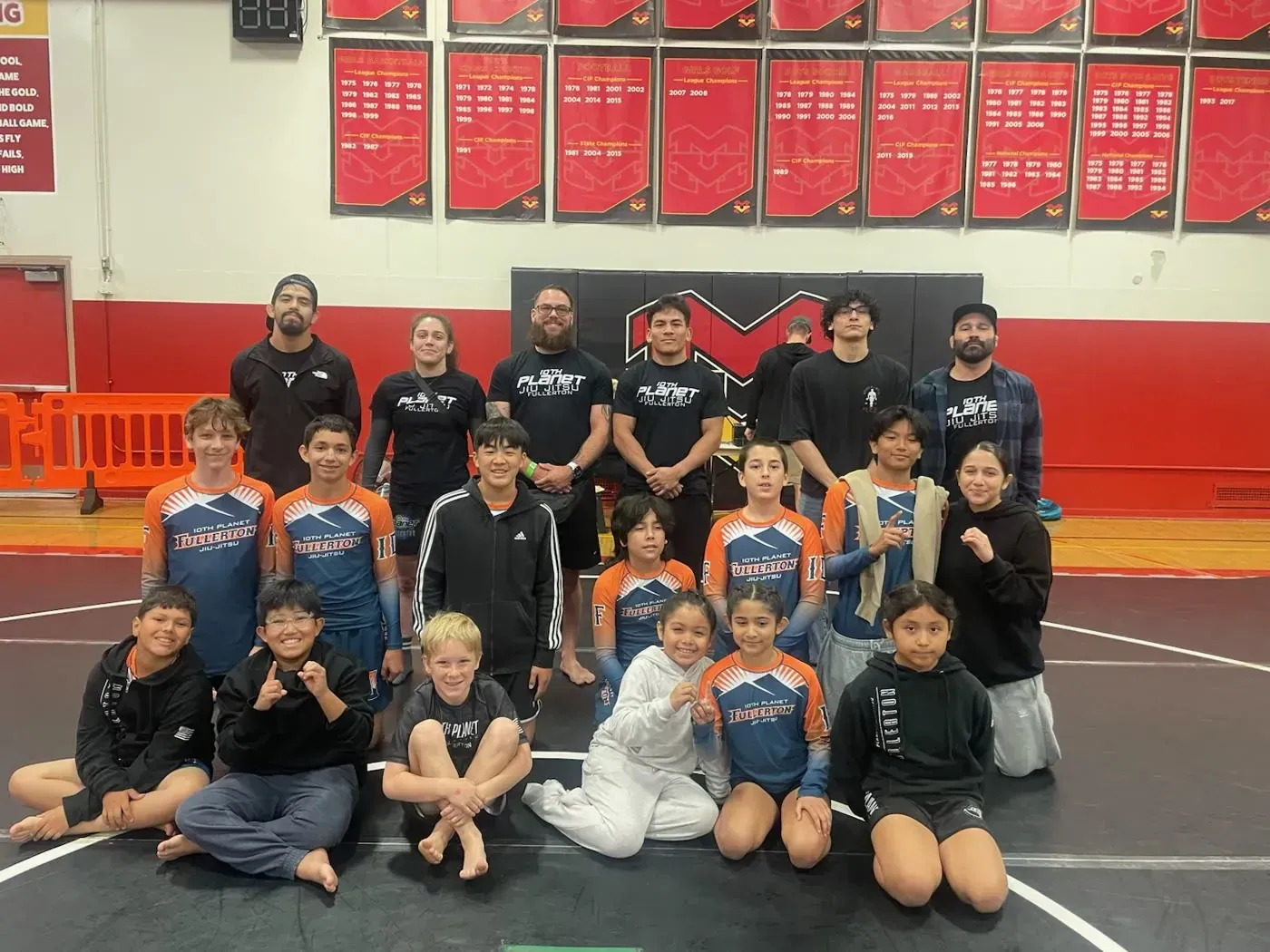 Youth wrestling team posing on gym mat in front of red wall banners, wearing black and orange uniforms.