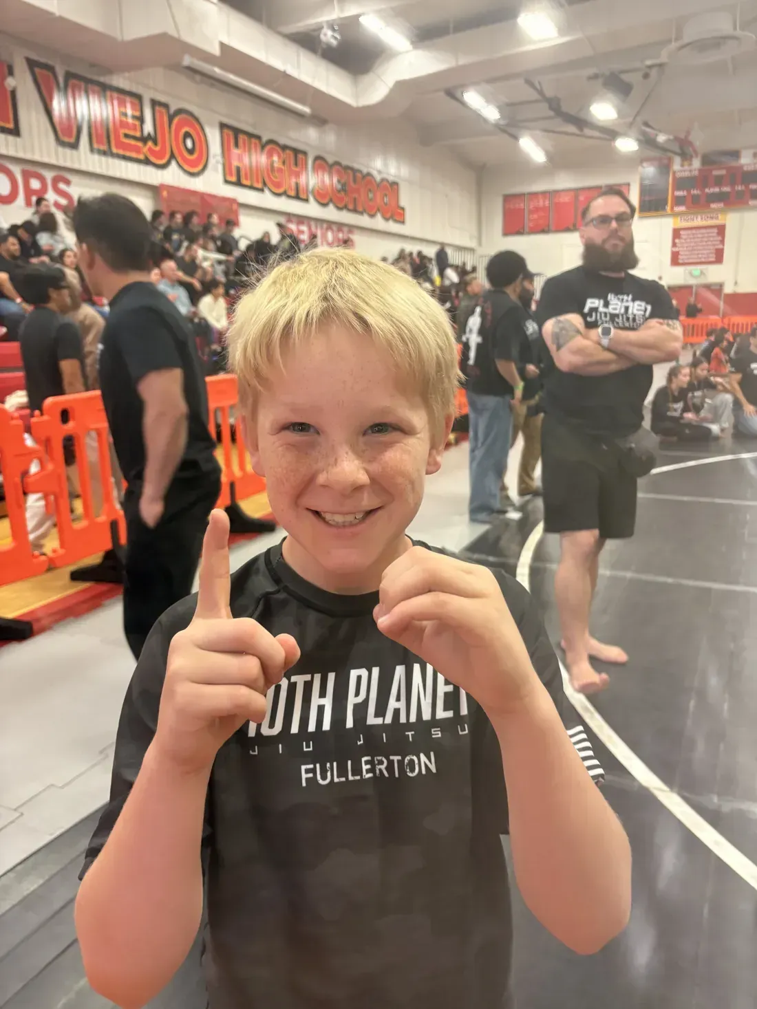 Smiling child in a gym, wearing a black “Youth Planet” shirt and holding up a fist pose.