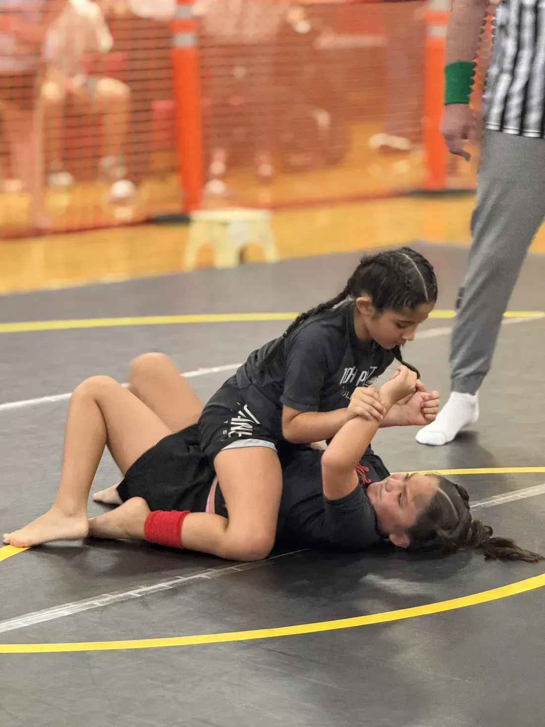 Two athletes grapple on a gym mat during a wrestling match, one pinning the other down.