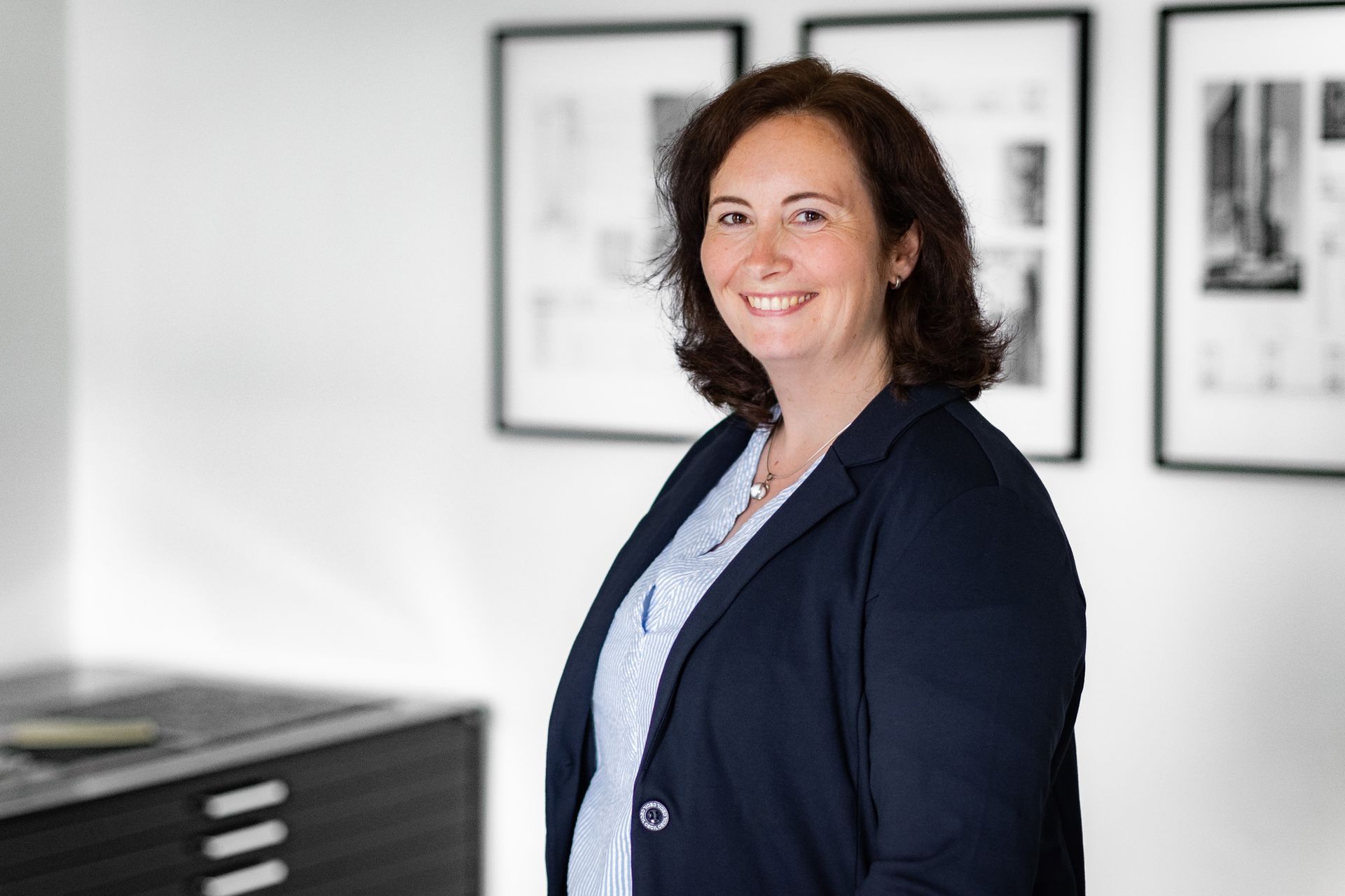 Eine Frau im Anzug lächelt vor einer Wand mit Bildern daran.