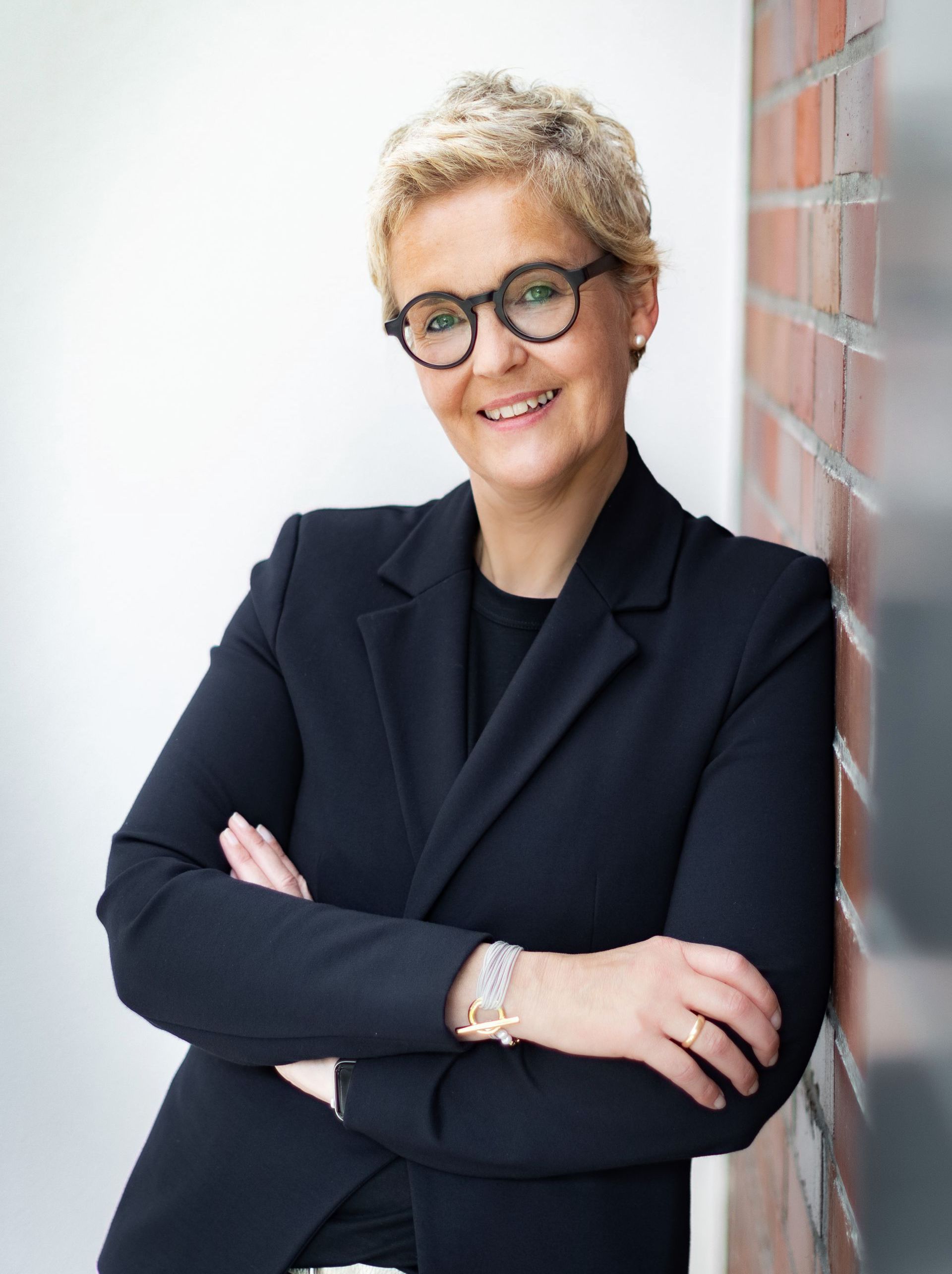 Eine Frau in Anzug und Brille lehnt an einer Backsteinmauer